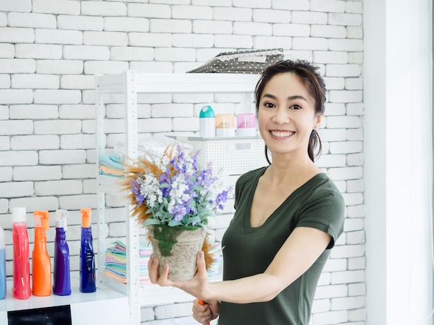 Bela jovem sorridente mulher asiática dona de casa usando o espanador de frango para espanar no vaso de flores na lavanderia.