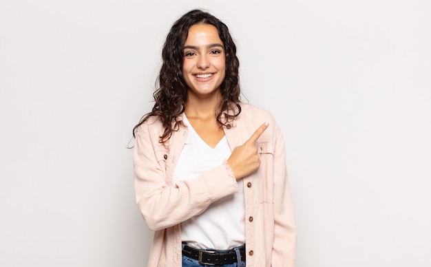 Bela jovem sorrindo alegremente, sentindo-se feliz e apontando para os lados e para cima, mostrando o objeto no espaço da cópia