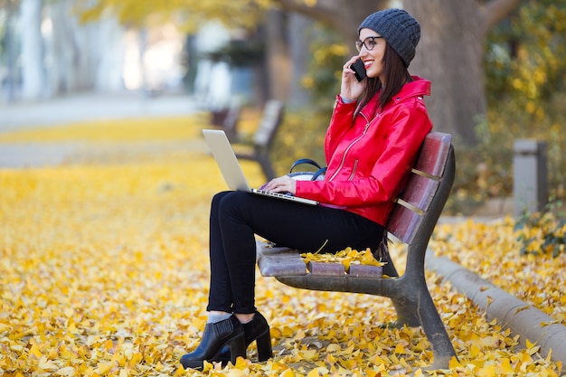 Bela jovem usando seu telefone celular enquanto trabalhava com laptop no outono.