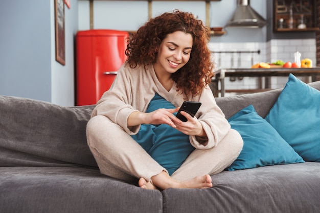 Foto bela jovem vestindo roupas de casa, sentada no sofá do apartamento e usando o celular