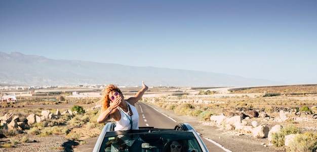 Foto bela jovem viajante manda um beijo ficando fora do teto de um carro conversível enquanto sua amiga dirige para uma viagem e férias no campo