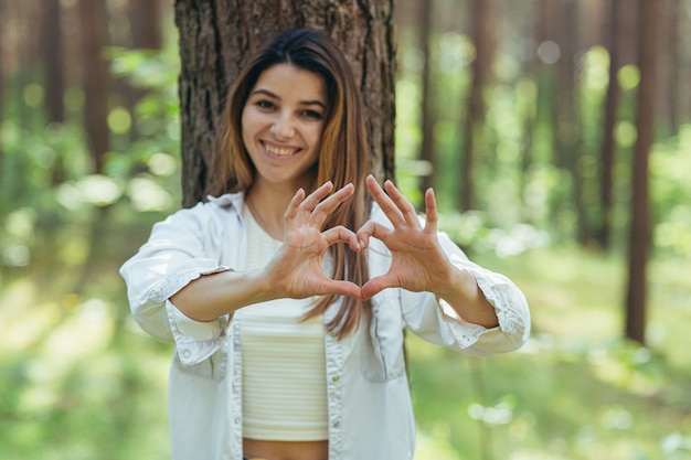 Bela jovem voluntária na floresta perto de uma árvore sorrindo olha para a câmera e mostra as mãos e os dedos um coração em sinal de amor pela natureza