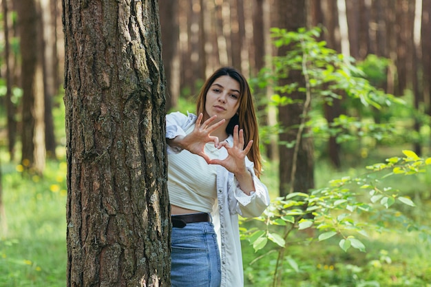 Bela jovem voluntária na floresta perto de uma árvore sorrindo olha para a câmera e mostra as mãos e os dedos um coração em sinal de amor pela natureza