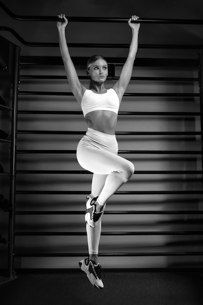 Bela loira alta se levanta na barra do ginásio contra o pano de fundo das barras da parede. Foto em preto e branco.