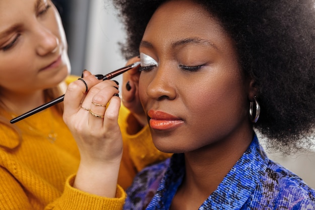 Bela maquiagem. Estilista muito jovem em um suéter amarelo grosso se sentindo inspirada ao colocar sombras prateadas nas pálpebras das modelos