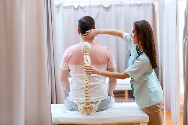 Bela médica segurando o modelo da espinha e examinando a espinha do paciente.