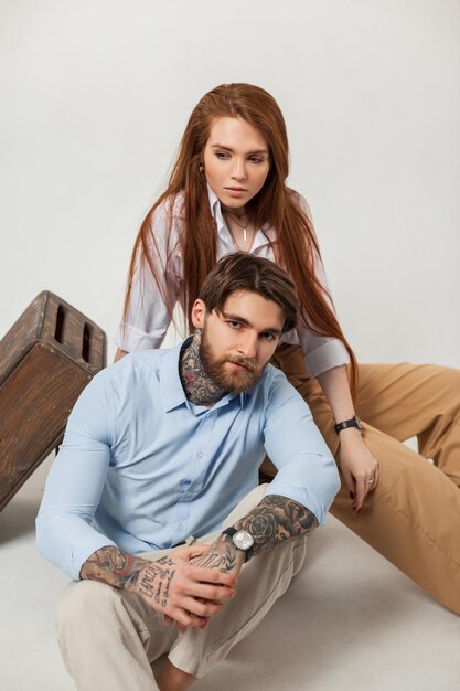 Bela moda casal ruiva linda mulher e elegante homem bonito brutal com barba e tatuagens em roupas da moda senta-se em um fundo branco no estúdio