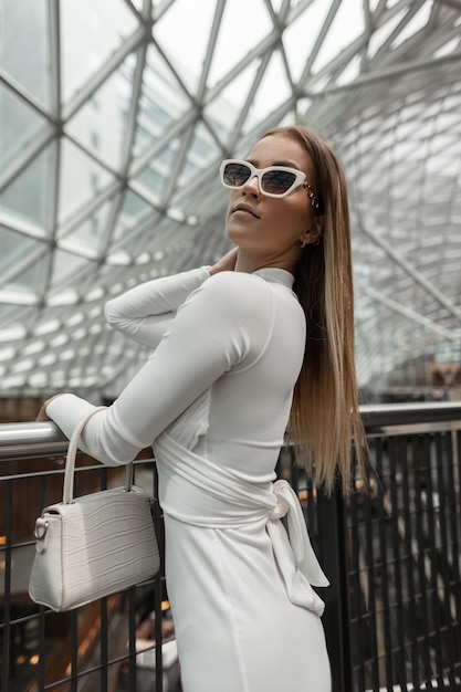 Bela modelo elegante jovem com óculos de sol brancos legais em um vestido branco elegante com uma bolsa de couro branco fica perto da grade de metal no shopping