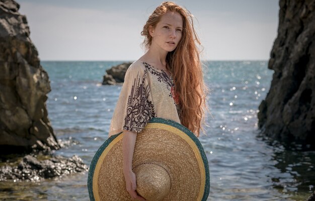 Bela modelo estilo boho vestindo blusa branca e sombrero posando na praia ao sol