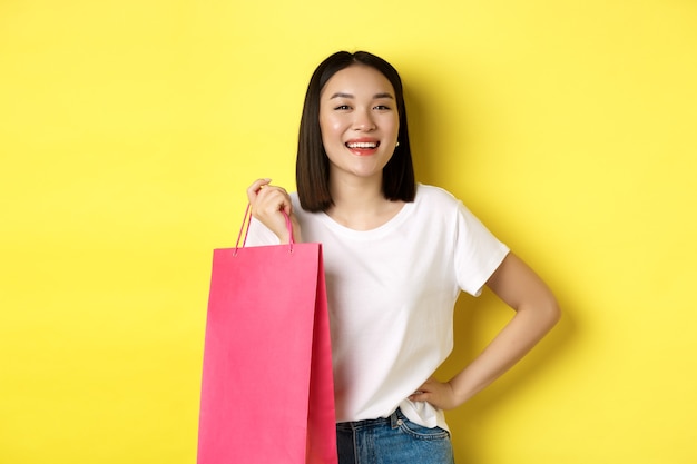 Bela modelo feminino asiático em t-shirt branca, segurando a sacola de compras e sorrindo satisfeito, em pé sobre um fundo amarelo.