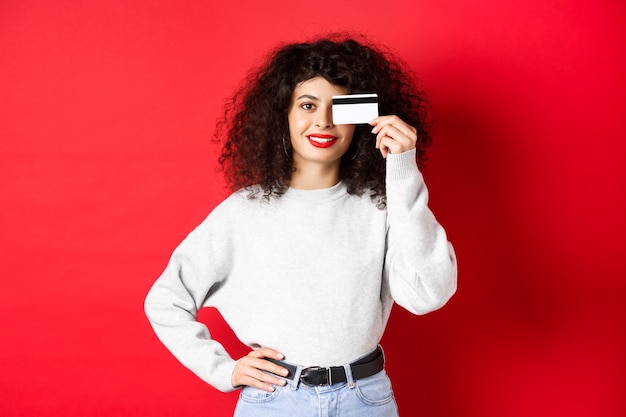 Bela modelo feminino com cabelo encaracolado, mostrando o cartão de crédito de plástico e sorrindo, em pé sobre um fundo vermelho. Conceito de maquiagem e compras