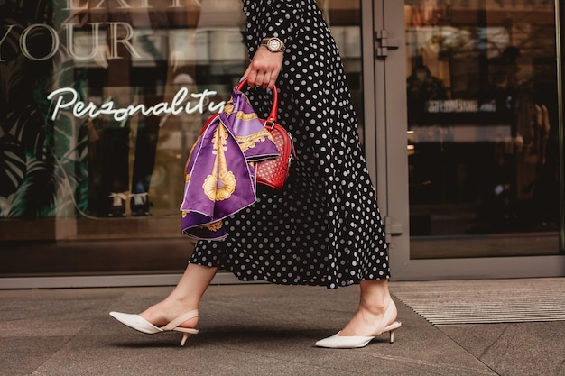 Bela modelo parece mulher morena com vestido branco com bolinhas pretas é ao acordar em um plano de fundo de rua da cidade com bolsas elegantes na mão.