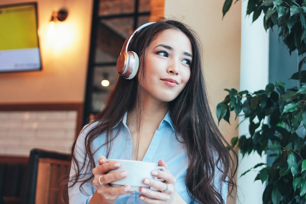 Bela morena encantadora sorridente menina asiática em fones de ouvido com uma xícara de café ou chá em goza de música no café