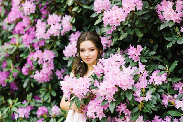 Bela morena jovem, com um sorriso e grandes olhos castanhos em jardins florescendo de rododendros rosa