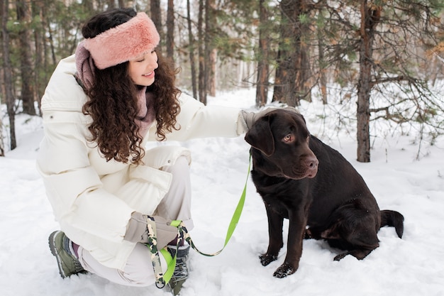 Bela morena jovem em trajes de inverno olhando para um retriever negro sentado na neve durante o frio em um parque ou floresta coberto de neve