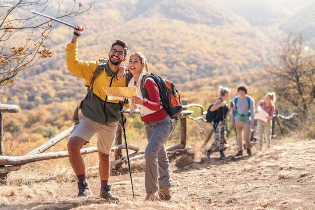 Bela morena sorridente segurando o mapa e olhando para o caminho certo enquanto homem apontando com vara. No fundo, o resto do grupo. Caminhadas na natureza no conceito de outono.