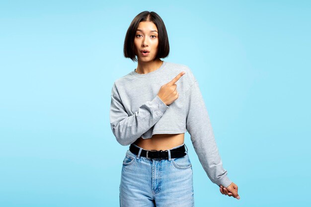 Foto bela mulher asiática sorridente vestindo roupas casuais apontando o dedo para o espaço de cópia olhando para a câmera de pé isolada em fundo azul conceito de compras de publicidade