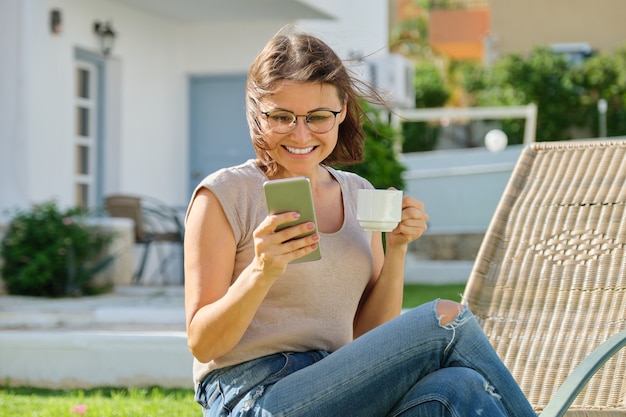 Bela mulher de meia idade descansando lendo, bebendo café conversando. Mulher sentada na espreguiçadeira do jardim no gramado perto da casa. Estilo de vida, lazer, tecnologia, beleza de pessoas maduras