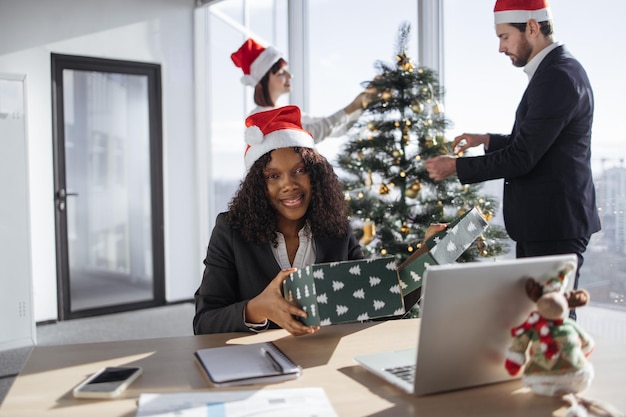 Bela mulher de negócios afro-americana sentada à mesa e abrindo um presente de Natal enquanto olha para a câmera no fundo de colegas de trabalho decorando a árvore de Natal