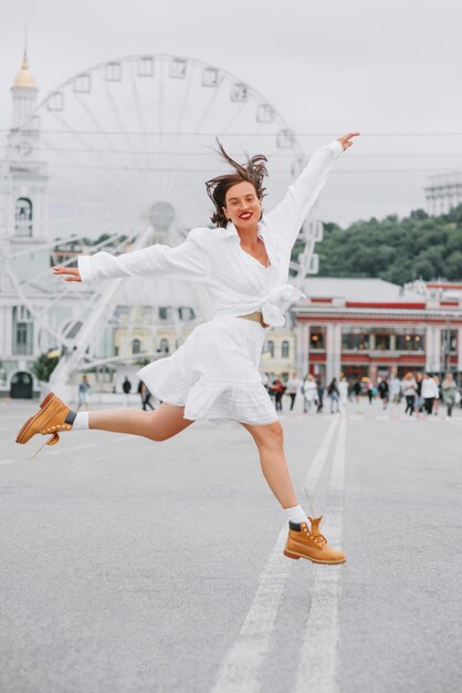 Bela mulher feliz é vestido branco está pulando na praça da cidade com a roda gigante na parte de trás