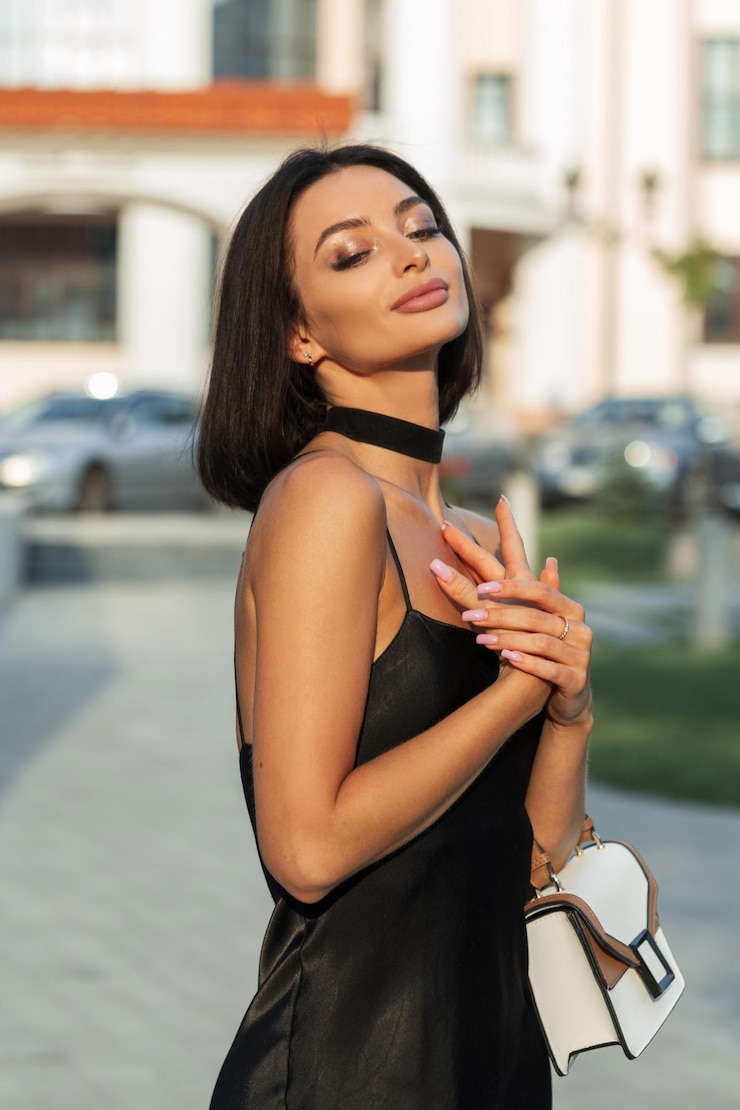 Foto bela mulher fresca sensual jovem com pele clara em um vestido preto à moda com tiras com uma bolsa elegante caminhando e desfrutando na cidade ao pôr do sol verão senhora bonita