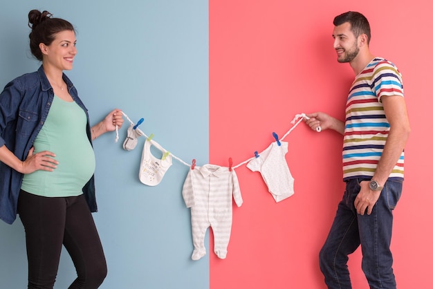 Bela mulher grávida e seu marido esperando bebê segurando bodysuits de bebê e sorrindo sobre fundo colorido