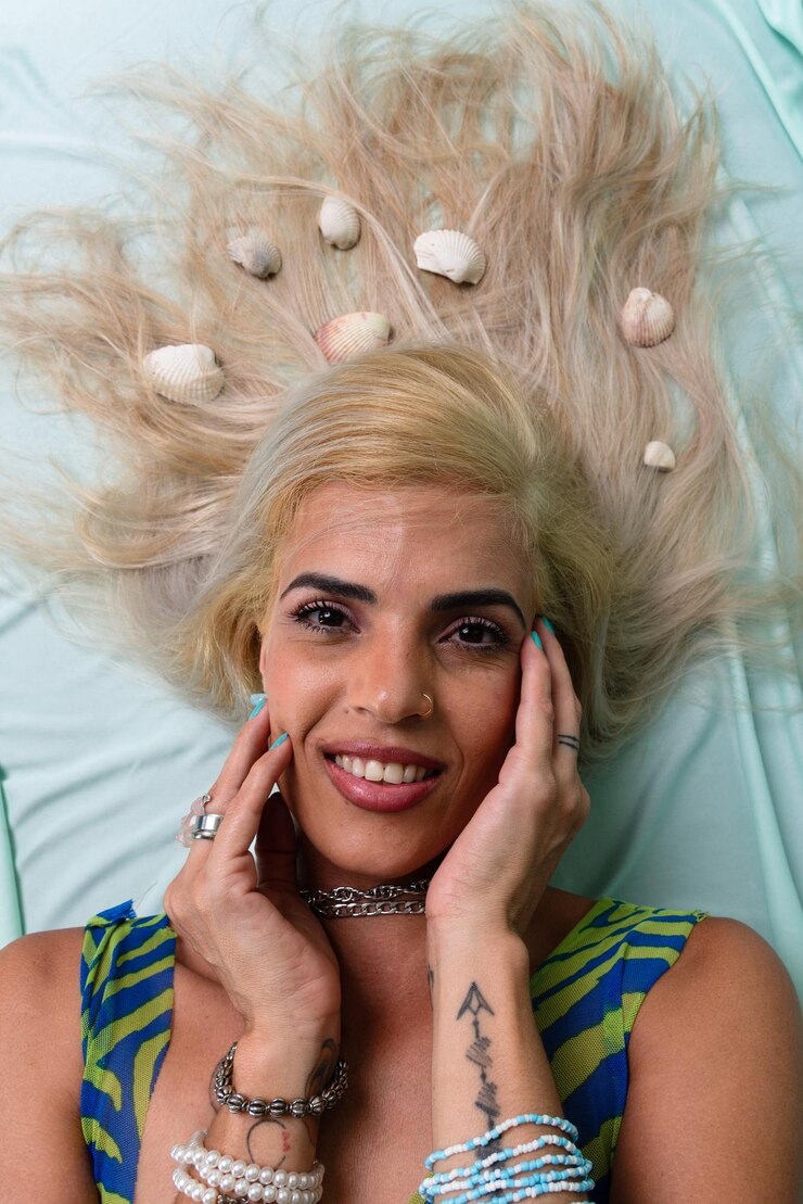 Foto bela mulher loira deitada em um sofá verde claro com conchas no cabelo olhando para a câmera