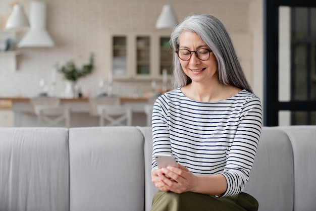 Bela mulher madura de meia-idade, mãe, esposa, freelancer, usando smartphone para verificar e-mails, redes sociais, trabalhando on-line em casa e fazendo compras.