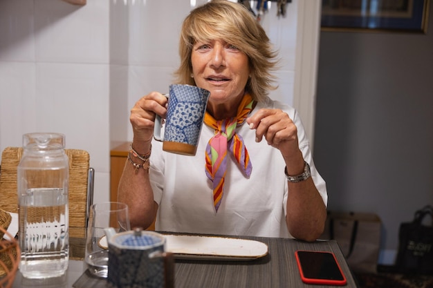 Foto bela mulher madura sorrindo olhando para a câmera tendo uma infusão na cozinha