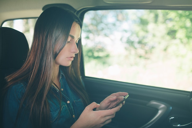 Bela mulher sentada nos bancos traseiros do passageiro no carro. Menina está usando um smartphone