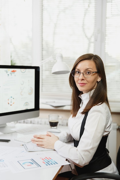 Foto bela mulher sorridente de cabelos castanhos de terno e óculos, sentada à mesa com uma xícara de café, trabalhando no computador com documentos em um escritório iluminado, olhando para a câmera