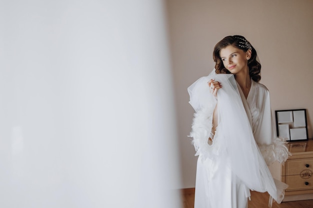 Foto bela noiva jovem com cabelos compridos e enrolados em roupa branca de moda perto da janela no hotel e segurando véu nas mãos bela jovem de manhã em um vestido branco espaço livre