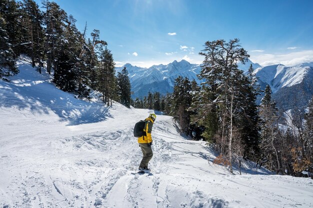 Bela paisagem da estância de esqui Arkhyz com floresta de neve de montanhas e homem snowboarder na jaqueta amarela e mochila em um dia ensolarado de inverno Montanhas do Cáucaso Rússia