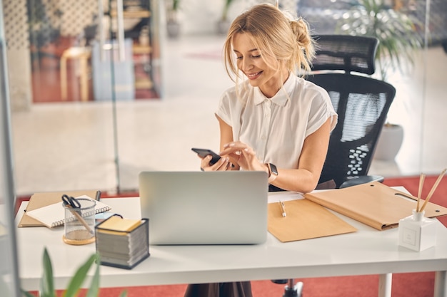 Bela trabalhadora sentada à mesa com o laptop e mensagens de texto no smartphone