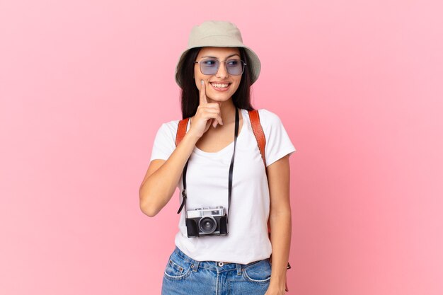 Bela turista hispânica sorrindo feliz e sonhando acordada ou duvidando com uma câmera fotográfica e um chapéu