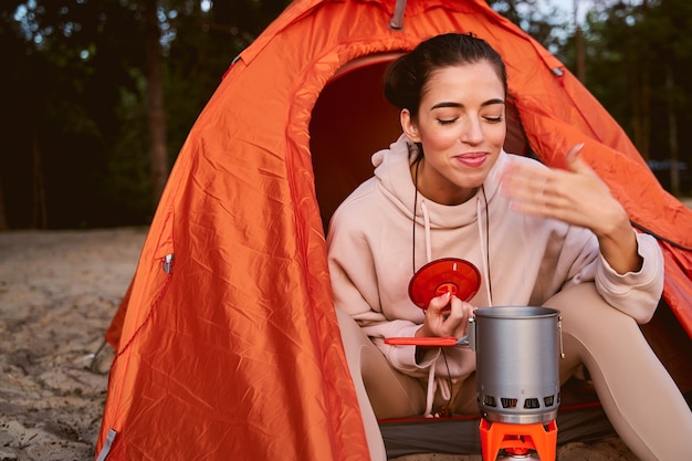 Bela viajante feminina de moletom preparando o café da manhã e sorrindo enquanto está sentada na barraca do acampamento e apreciando o cheiro da bebida