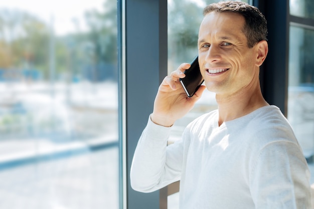 Bela Vista. Homem bonito e positivo em pé perto da janela e conversando por telefone enquanto olha para você