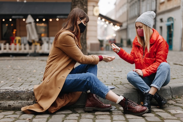 Belas moças em roupas quentes, sentado na rua da cidade e usando desinfetante. Mulheres bonitas usando máscaras protetoras enquanto passam algum tempo juntas.