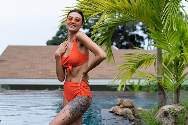 Belas mulheres europeias em biquíni laranja de duas peças em perfeita forma, bom corpo bronzeado com óculos escuros da moda ao ar livre na piscina tropical e palmeira