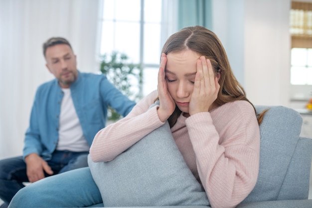 Beleidigte Tochter, die Hände in der Nähe ihres Gesichts hält und sich von Vater abwendet, der neben ihm auf der Couch sitzt.