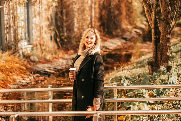 Beleuchtet von goldenem, warmem Licht, steht eine wunderschöne Blondine mit einer Tasse heißen Kaffees in ihren Händen im Herbststadtpark auf einer Brücke über einem Bach, der sich in ein helles Band verwandelt hat. Fabelhafte Herbstlandschaft.