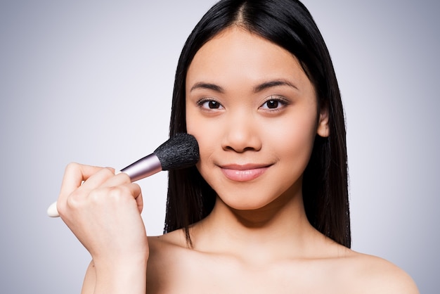 Beleza corada. Mulher asiática jovem e bonita sem camisa segurando um pincel de maquiagem na bochecha e sorrindo em pé contra um fundo cinza