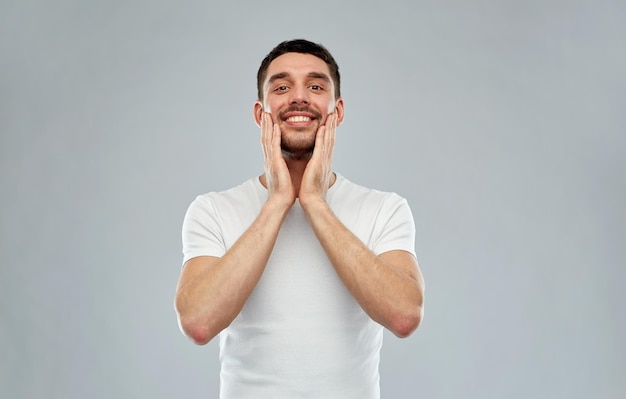 beleza, cuidados com a pele, aliciamento e conceito de pessoas - jovem feliz tocando seu rosto aplicando loção pós-barba sobre fundo cinza