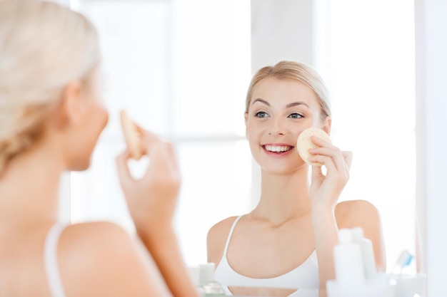 beleza, cuidados com a pele e conceito de pessoas - jovem sorridente lavando o rosto com esponja de limpeza facial no banheiro