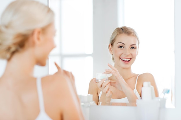 Beleza, cuidados com a pele e conceito dos povos - jovem mulher de sorriso que aplica o creme para enfrentar e que olha para espelhar em casa o banheiro