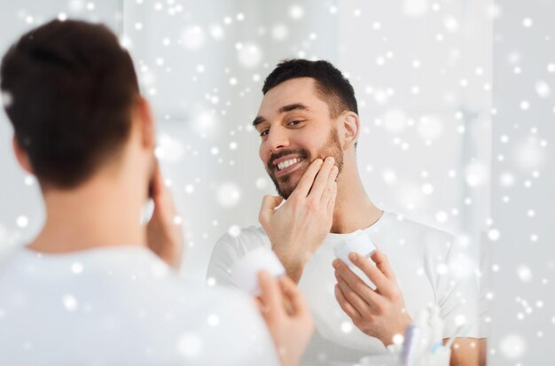 beleza, cuidados com a pele, inverno e conceito de pessoas - jovem sorridente aplicando creme no rosto e olhando para o espelho no banheiro de casa sobre a neve