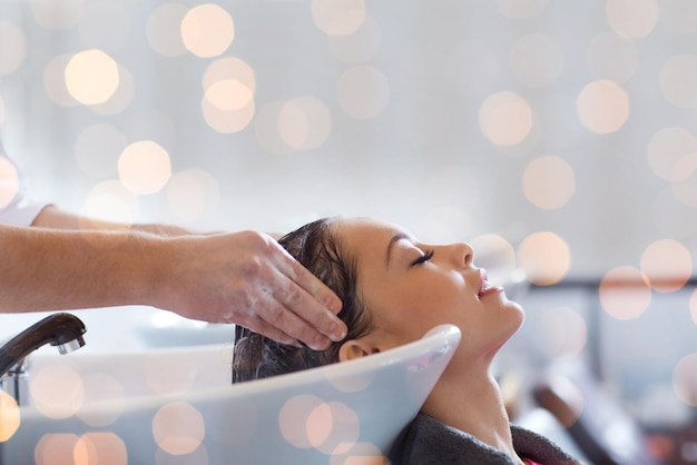 Foto beleza, cuidados com os cabelos e conceito de pessoas - jovem feliz com cabeleireiro lavando a cabeça no salão de cabeleireiro sobre luzes de férias