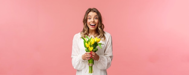 Beleza de férias e conceito de primavera retrato de jovem loira linda animada e espantada em vestido branco segurando tulipas amarelas receber flores como presente smling divertido fundo rosa