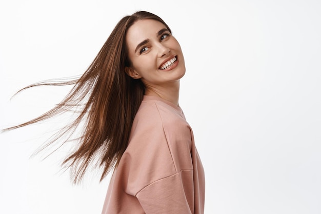 Beleza e estilo de vida. Mulher jovem e atraente feliz com cabelo natural e saudável, penteado voando no ar, garota sincera sorri e se vira para trás, olhando de lado para o logotipo, fundo branco