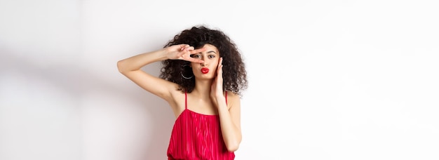 Foto beleza e moda elegante mulher de cabelos encaracolados em vestido vermelho mostrando vsign nos olhos e lábios franzidos